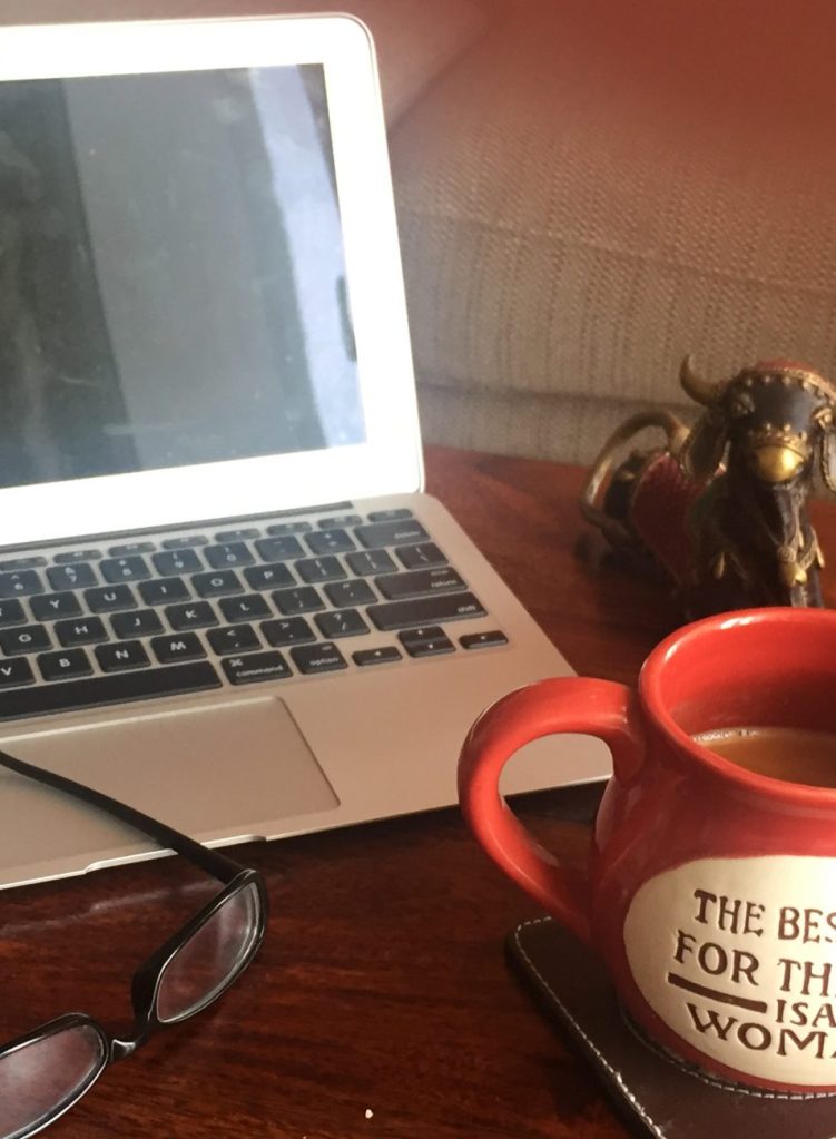 A workspace featuring a laptop, a red coffee mug with an inspiring quote, reading glasses, and a decorative figurine, all set on a wooden table.